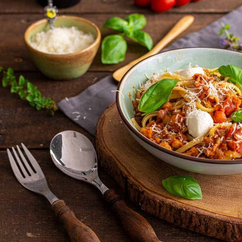 Nahaufnahme einer dampfenden Linsenbolognese mit roten Linsen auf Spaghetti, serviert in einer weißen Schale mit frischen Kräutern